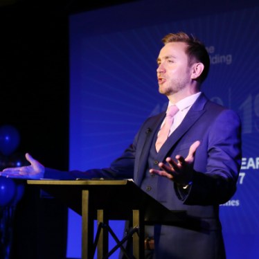 Kevin Durham presenting from a lectern on a blue-lit stage at a corporate conference awards evening