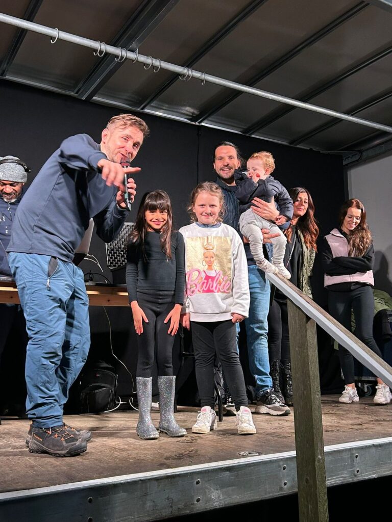 Kevin Durham on stage at a family event with two young girls, a father holding a baby and other relatives waiting to play