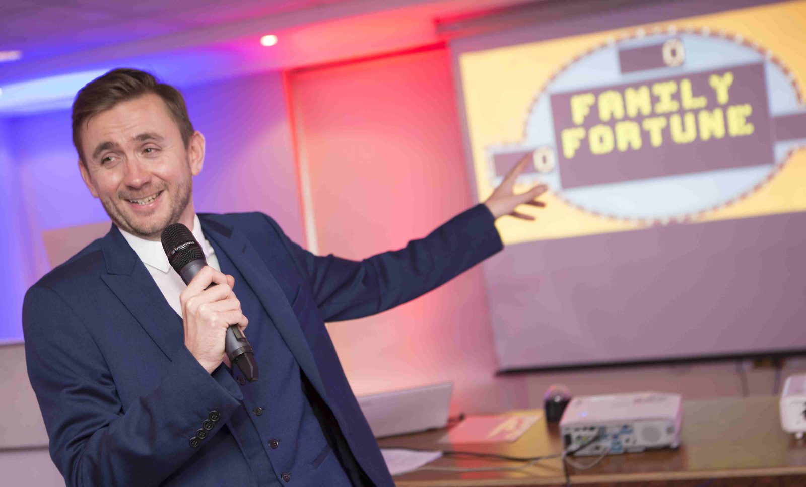 Kevin Durham hosting a Family Fortunes style game show, microphone in hand, presenting the board to a corporate audience