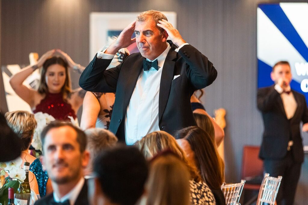 A guest in black tie with his hands on his head during Heads or Tails, Kevin Durham on the microphone behind him